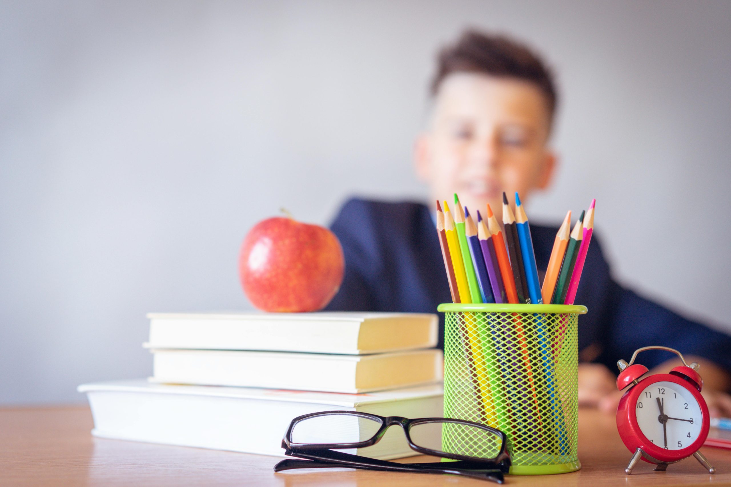 Un banco di scuola con libri, matite colorate, occhiali e una mela, in lontananza un bambino seduto.